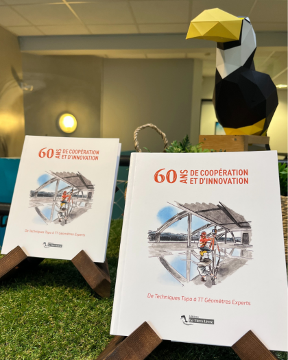 Deux livres retraçant l'histoire de TT Géomètres Experts posé sur des supports en bois sur une table recouverte de pelouse artificielle. Un toucan de décoration est à l'arrière-plan.